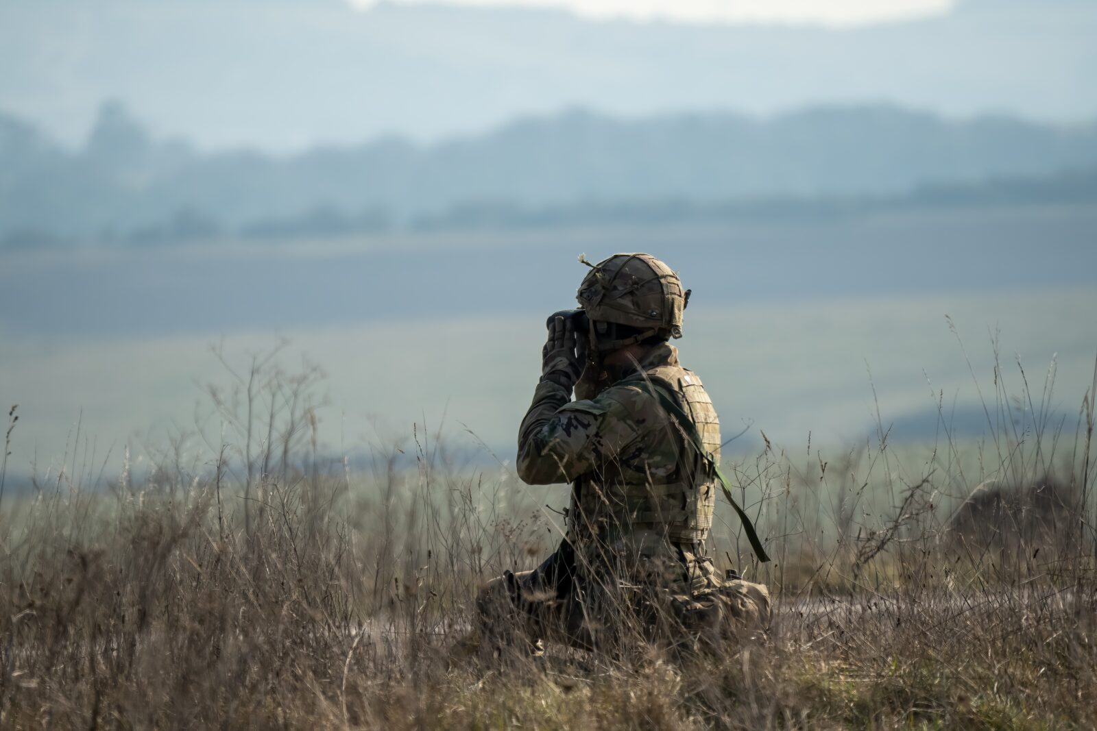 A British army infantry soldier using binoculars to scan distance for enemy threats on a military exercise in Wiltshire