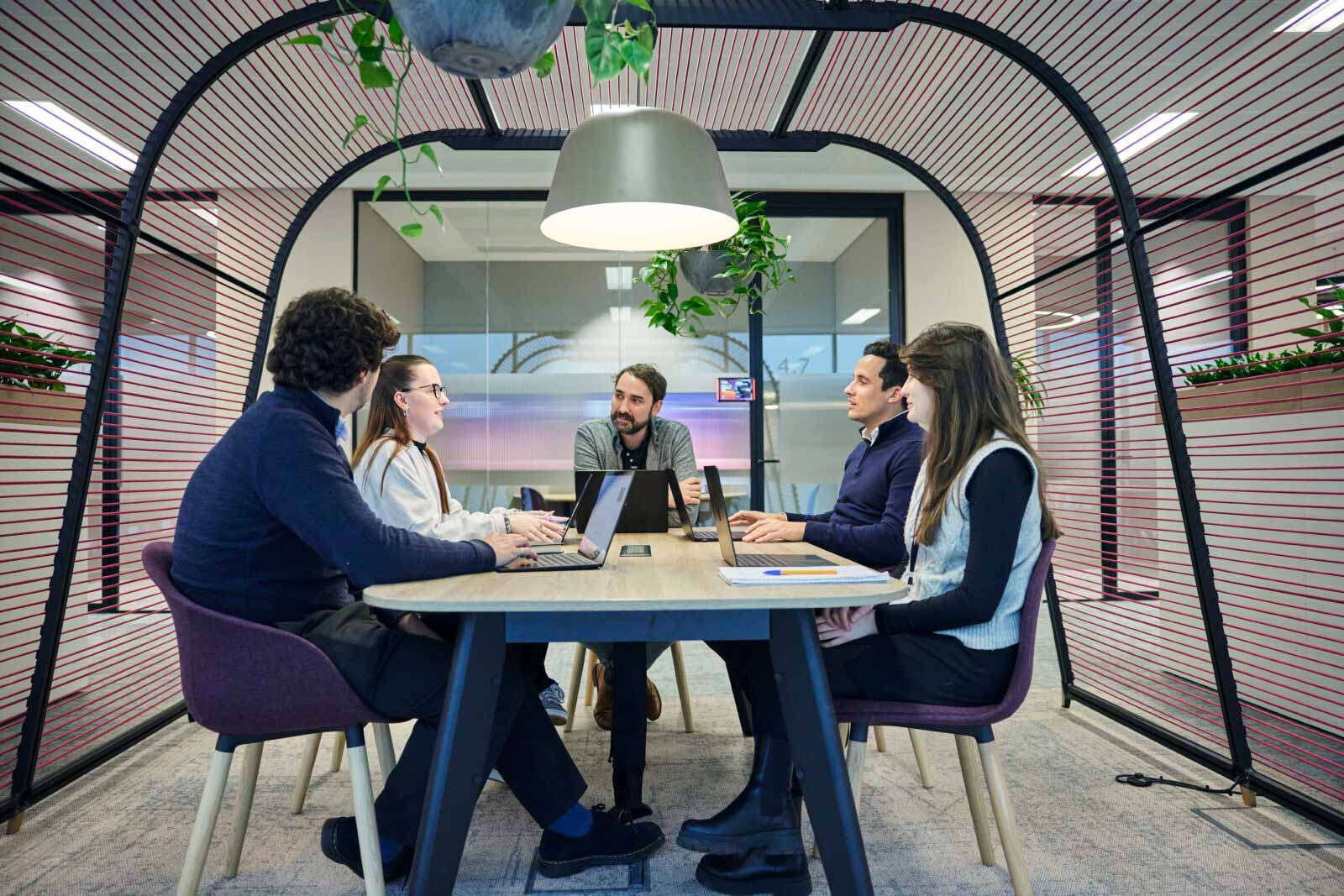 Five Burges Salmon employees in a modern office pod space, sitting around a table in discussion and using their laptops
