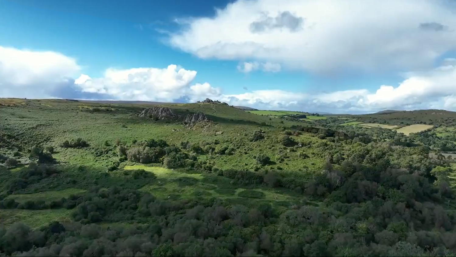 Thumbnail image of a slow moving Drone Shot over a green field and hillside with blue sky and white clouds in the background