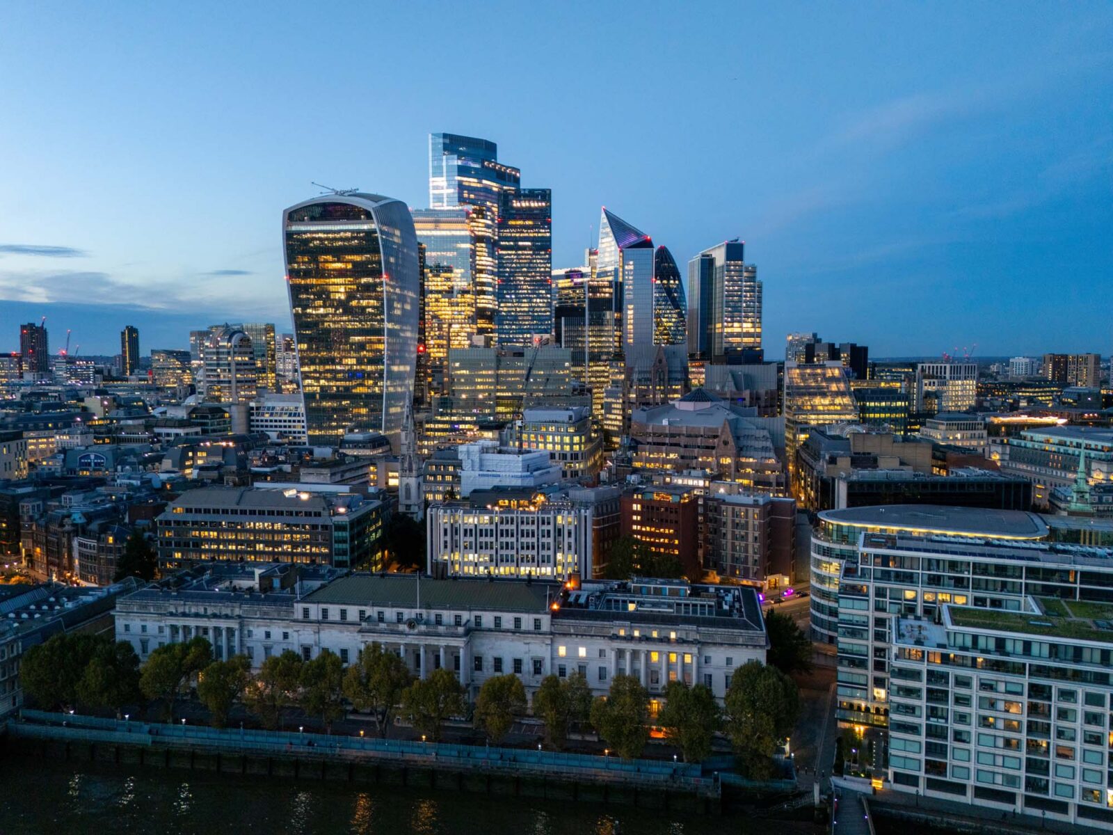 City of London skyline at dusk with illuminated office buildings beside the River Thames.
