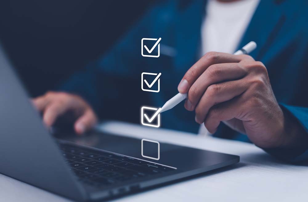 Person using a stylus next to a laptop with a digital checklist showing three completed tick boxes and one remaining item